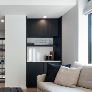 North Vancouver Living Room Renovation with Custom Built Bar Cabinets North Vancouver stylish built-in bar area featuring matte black cabinetry, a white countertop, mirrored backsplash, glassware display shelf, and subtle recessed lighting, nestled beside a modern living room with a neutral-toned sofa.