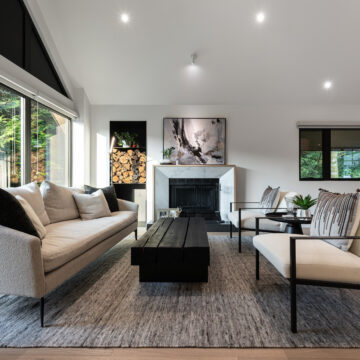 Modern Living Room with Wood Storage Feature and Natural Light Contemporary living room with vaulted ceilings, a neutral sofa, black wood coffee table, built-in firewood storage with integrated lighting, a black fireplace, and large windows bringing in natural light.