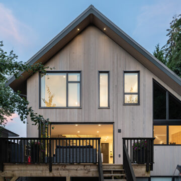 North Vancouver Home Renovation Back Exterior with Large Windows and Deck Rear view of a modern home at dusk featuring vertical light-toned siding, black-framed windows, a sloped roof, and a spacious raised deck with black railings, warmly lit from within and surrounded by mature trees.