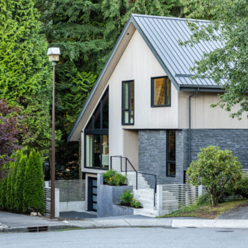 Modern Sloped-Roof Renovation Nestled in North Vancouver Forest Setting Street view of a contemporary home in North Vancouver with a steep metal roof, vertical siding, dark stone accents, and black-framed windows, surrounded by mature trees and lush landscaping.