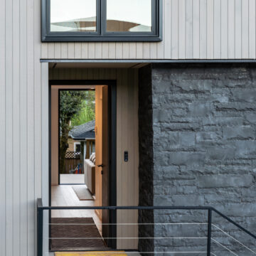 North Vancouver Modern Front Entry with Vertical Siding and Stone Wall Detail Architectural detail of a modern home entryway featuring vertical light-toned wood siding, a textured dark stone wall, a sleek glass door framed in black metal, and a view through the foyer to the backyard.