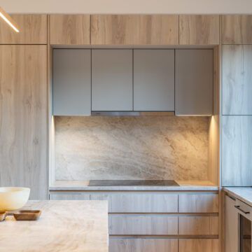North Vancouver Kitchen Renovation Close-Up of Cabinetry with Stone Backsplash Detail view of a modern kitchen featuring light wood cabinetry, matte grey upper cabinets, integrated cooktop, and a full-height stone slab backsplash with under-cabinet lighting for a clean, upscale look.