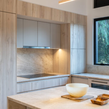Custom Light Wood Kitchen with Stone Surfaces and Picture Window Modern kitchen design featuring light wood cabinetry, a full stone slab backsplash, built-in cooktop, and a large window with forest views, complemented by a minimalist island with neutral decor.