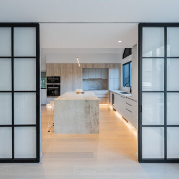 North Vancouver Minimalist Kitchen Renovation Bright modern kitchen with a large center island, light wood cabinetry, under-cabinet lighting, and frosted glass sliding doors framed in black metal, showcasing clean lines and contemporary design.