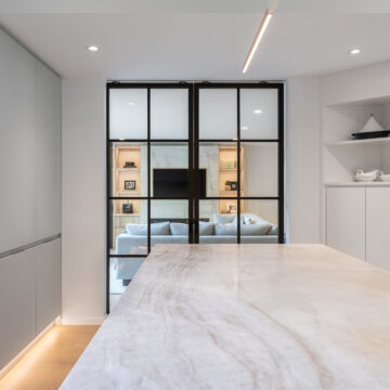 North Vancouver Kitchen Renovation with Engineered Stone Island and Black-Framed Glass Doors Contemporary kitchen island with polished quartz surface, facing black-framed glass doors that open to a cozy living room with built-in shelving and soft lighting, surrounded by sleek cabinetry and integrated lighting.