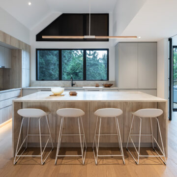 North Vancouver Kitchen Renovation with Vaulted Ceiling and Oversized Island Seating  Integrated Refrigerator and Modern White Stools for Island Contemporary kitchen featuring a large central island with four white bar stools, vaulted ceiling with black clerestory windows, minimalist cabinetry, and large black-framed windows bringing in natural forest views.