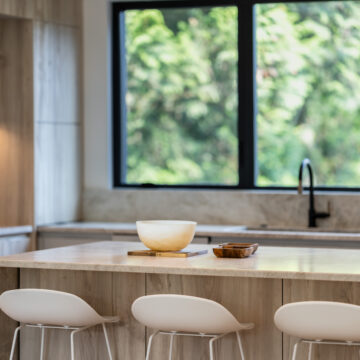 Kitchen Island Seating with Forest Views and Natural Light Close-up of a modern kitchen island with light wood cabinetry, white bar stools, a stone countertop, and a matte black faucet set against large windows framing lush green forest views.