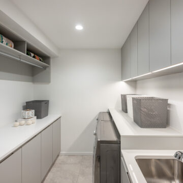 North Vancouver Streamlined Laundry Room Renovation Modern laundry room featuring sleek grey cabinetry, quartz countertops, built-in washer and dryer, undermount sink, open shelving, and minimalist design with under-cabinet lighting for a clean, functional workspace.