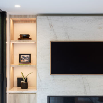North Vancouver Custom Millwork and Stone Fireplace Surround Close-up of a modern living room fireplace clad in marble-look stone, flanked by built-in illuminated display shelves styled with framed art, books, and plants, and a wall-mounted flat screen TV above the firebox.
