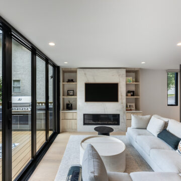 North Vancouver Scan-Design Inspired Family Room Elegant family room renovation featuring a sleek gas fireplace with built-in shelving, wall-mounted TV, and full-height sliding glass doors opening to an outdoor patio, complemented by a light-toned sectional and nested coffee tables.