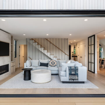 North Vancouver Family Room Renovation with Sliding Glass Wall Wide view of a stylish open-concept living room with a large sliding glass wall opening to a patio, featuring a light-toned sectional, layered coffee tables, a modern fireplace with TV, and a striking vertical slat wood staircase backdrop.