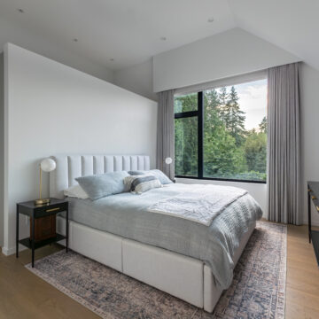 North Vancouver Primary Room Renovation with Large Window North Vancouver tranquil primary bedroom featuring a tufted upholstered bed, soft grey bedding, black nightstands, a vintage-style rug, and a large black-framed window showcasing forest views, all under a vaulted ceiling with recessed lighting.