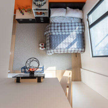 North Vancouver Kid's Bedroom Renovation from Loft Bird's-eye view of a bedroom renovation featuring a plaid bed, orange accent wall, mini basketball hoop, sports-themed decor, and a built-in loft with wooden ladder, showcasing a bright and functional play-and-rest space.
