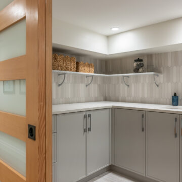 Custom laundry room featuring soft grey built-in cabinetry, open shelving, a white quartz countertop, patterned vertical tile backsplash, and wood shaker sliding doors.