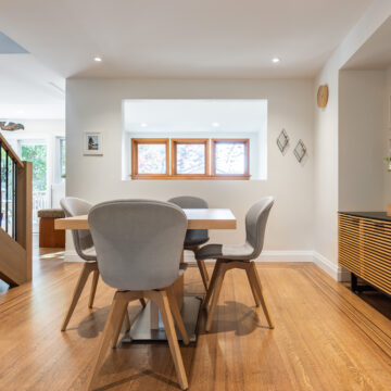 Renovated dining area featuring a wood dining table, upholstered chairs, light wood flooring, and a modern staircase with wood treads and black metal railing, creating an open and cohesive layout.