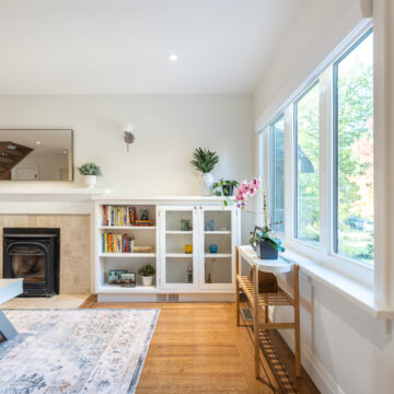 Bright living room renovation featuring a classic fireplace with tiled surround, custom white built-in shelving and cabinetry, large picture windows, and warm wood flooring styled with layered textiles and plants.