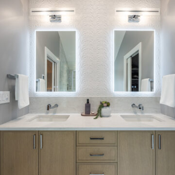 Vancouver, Custom primary ensuite double vanity with wood cabinetry, integrated stone countertop, dual undermount sinks, wall-mounted faucets, and backlit mirrors set against a textured geometric tile feature wall.