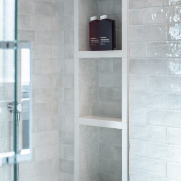 Custom shower niche with integrated shelving, framed in white stone and set within light gray rectangular tile, part of a modern bathroom renovation with glass shower enclosure.