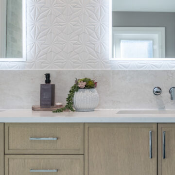 Custom double bathroom vanity with light wood cabinetry, integrated LED mirrors, geometric textured tile backsplash, quartz countertop, and wall-mounted chrome faucets in a modern ensuite renovation.
