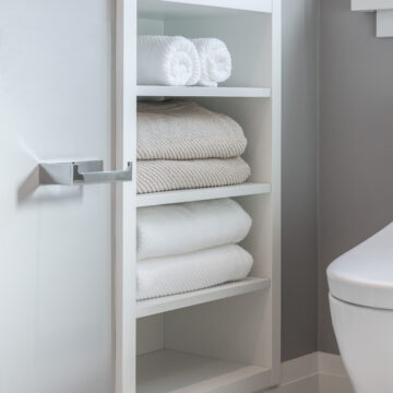 Custom bathroom linen niche with built-in white shelving, neatly stacked towels, hex tile flooring, and modern chrome hardware in a minimalist bathroom renovation.