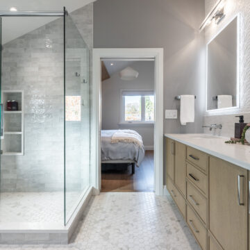 Bright primary ensuite featuring a frameless glass shower with built-in niche, light stone tile walls, hex tile flooring, and a custom wood vanity with integrated lighting and textured feature tile.