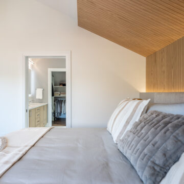 Primary bedroom featuring a sloped wood-clad ceiling, custom upholstered headboard, layered neutral bedding, and direct access to a walk-in closet and ensuite bathroom.