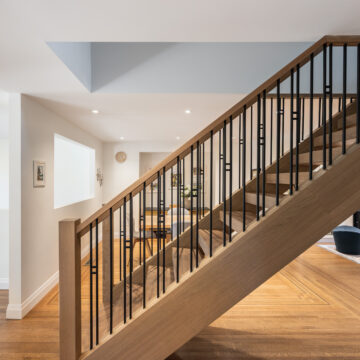 Open-concept interior featuring a custom oak staircase with black metal balusters, warm wood flooring, and sightlines into a modern living and dining area.